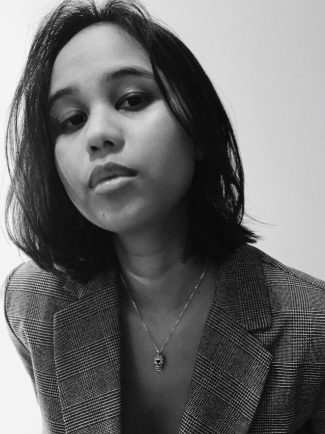 Black-and-white portrait of Keziah Abigail, looking at the camera, with dark shoulder-length hair, a checked blazer and a small pendant necklace.